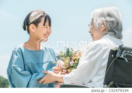 車椅子に乗るおばあちゃんに花束をプレゼントする笑顔の孫娘 車椅子に乗るおばあちゃんに花束をプレゼントする笑顔の孫娘 132479194