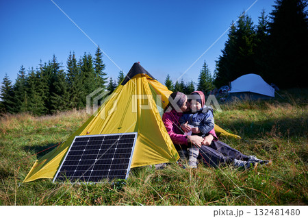 Young child and mother play with smartphone while charging with photovoltaic solar panel near tourist tent in summer. Integration of renewable energy in outdoor camping activities. 132481480
