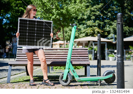 Happy woman charging her electric scooter from solar panel. Young female holding solar battery and catching sunlight. Concept of renewable energy resources. Happy woman charging her electric scooter from solar panel. Young female holding solar battery and catching sunlight. Concept of renewable energy resources. 132481494