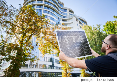 Man holding photovoltaic solar panel in front of new building. Concept of integration of sustainable renewable energy sources into modern architecture. 132481500