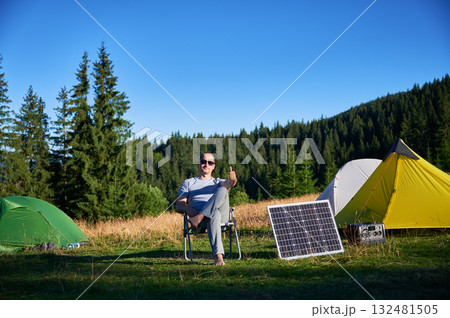 Man using photovoltaic solar panel for charging portable power station outdoor in eco-friendly camping. Male tourist giving thumbs up near tents. On background tall pine trees and clear blue sky. Man using photovoltaic solar panel for charging portable power station outdoor in eco-friendly camping. Male tourist giving thumbs up near tents. On background tall pine trees and clear blue sky. 132481505