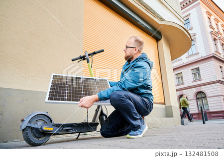 Male person holds photovoltaic solar panel, for charging electric scooter in urban settings. Integration of solar power as sustainable energy source, promoting eco-friendly urban transportation. Male person holds photovoltaic solar panel, for charging electric scooter in urban settings. Integration of solar power as sustainable energy source, promoting eco-friendly urban transportation. 132481508