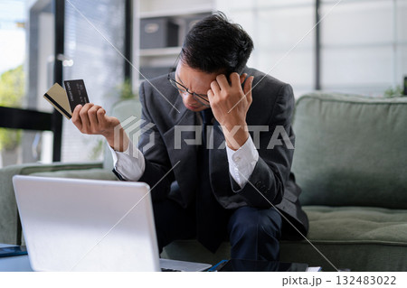 Asian man stressed over credit card debt, holding bills in frustration. Financial anxiety and economic burden concept in a realistic lifestyle setting 132483022