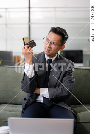 Asian man stressed over credit card debt, holding bills in frustration. Financial anxiety and economic burden concept in a realistic lifestyle setting Asian man stressed over credit card debt, holding bills in frustration. Financial anxiety and economic burden concept in a realistic lifestyle setting 132483030