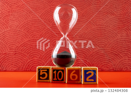 Hourglass with black sand with a wooden number block on a red background. Time passing or time flowing. Sand glass, sand timer, sand clock. Concept of time 132484308