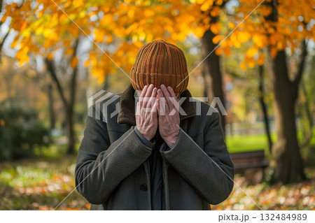 An elderly man stands in a park, covering his face with both hands, while colorful autumn leaves fall around him 132484989