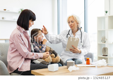 A doctor checks a young girl's temperature while her mother comforts her in a doctor's office, with a teddy bear. A doctor checks a young girl's temperature while her mother comforts her in a doctor's office, with a teddy bear. 132485127