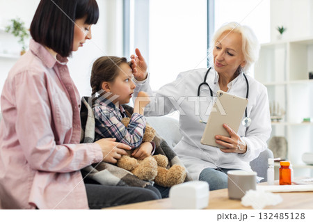 A young girl with a cold is being examined by a doctor while her mother comforts her. The doctor is checking the child's temperature with a tablet in hand. 132485128