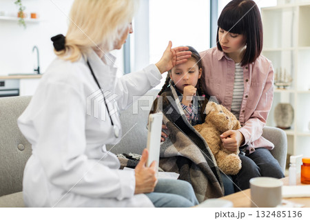 A concerned doctor checks a young girl's temperature while the mother comforts her. The child is wrapped in a blanket and holding a teddy bear. 132485136