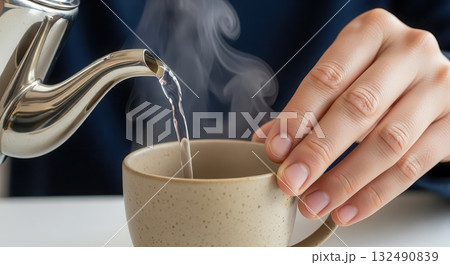 Hand pouring steaming water from a kettle into a ceramic cup, showcasing the brewing process, with gentle steam rising, creating a warm and inviting atmosphere for tea or coffee lovers Hand pouring steaming water from a kettle into a ceramic cup, showcasing the brewing process, with gentle steam rising, creating a warm and inviting atmosphere for tea or coffee lovers 132490839