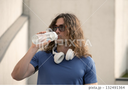 Handsome sporty fit athlete drinking water from bottle in sunny day against urban white wall. Water balance. Handsome sporty fit athlete drinking water from bottle in sunny day against urban white wall. Water balance. 132491364