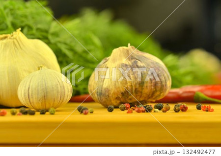 A bulb of garlic, red chili peppers, four-color pepper, and carrot tops on a wooden cutting board. The concept of cooking and vegetables in the home. Garlic - Allium sativum is a species of bulbous A bulb of garlic, red chili peppers, four-color pepper, and carrot tops on a wooden cutting board. The concept of cooking and vegetables in the home. Garlic - Allium sativum is a species of bulbous 132492477