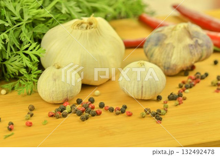 A bulb of garlic, red chili peppers, four-color pepper, and carrot tops on a wooden cutting board. The concept of cooking and vegetables in the home. Macro image, close-up. Garlic - Allium sativum is 132492478