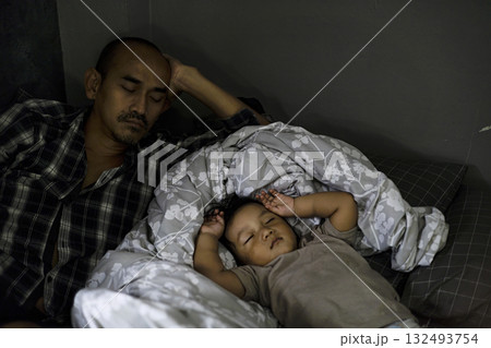 Dad Sleeping Beside Young Child In Daytime, A peaceful dad naps beside a young child, soft sunlight warming the cozy scene, perfect for a family photography shot or a tender bonding showcase. 132493754