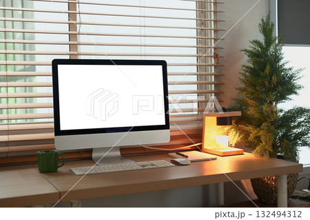 Minimalist workspace with computer, desk lamp, and coffee mug by window in warm light. 132494312