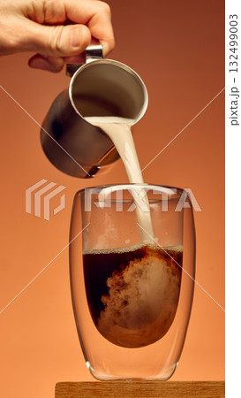 Milk poured from metal pitcher into black coffee in transparent glass showing creamy swirl Milk poured from metal pitcher into black coffee in transparent glass showing creamy swirl 132499003