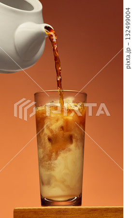 Coffee poured from white pot into tall glass creating swirl pattern over brown background 132499004