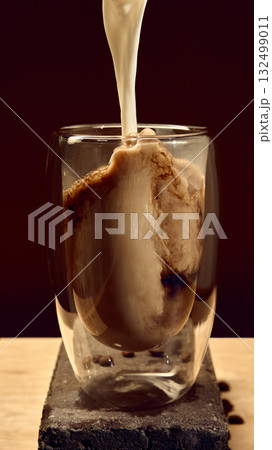 Milk pouring into glass of dark coffee with swirling texture and roasted beans on stone block Milk pouring into glass of dark coffee with swirling texture and roasted beans on stone block 132499011