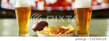 Super Bowl game party, glass of beer and plate of chips with salsa next to television screen 132500128