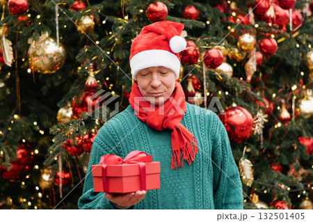 Portrait of happy man wearing green sweater and Santa Hat holding red gift box near Christmas tree. Xmas Holiday. Close up. 132501405