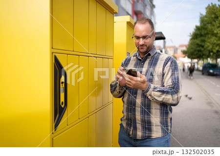 Man using smartphone while standing near bright yellow delivery lockers in an urban environment during daytime 132502035