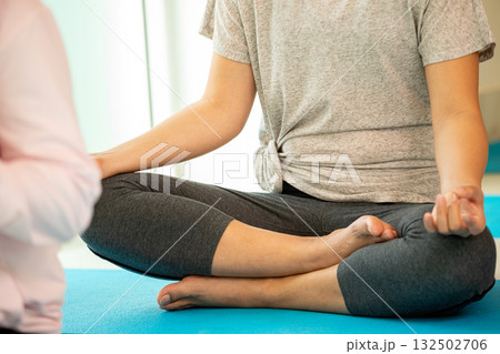 Close-up of a woman sitting cross-legged in lotus pose on a blue yoga mat, meditating indoors with hands in mudra. Concept of mindfulness, inner peace, yoga practice, and stress relief Close-up of a woman sitting cross-legged in lotus pose on a blue yoga mat, meditating indoors with hands in mudra. Concept of mindfulness, inner peace, yoga practice, and stress relief 132502706