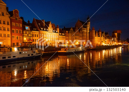 Gdansk riverside at night showcases historic buildings beautifully lit, reflecting on Motlawa River. People stroll along the promenade, enjoying a peaceful evening atmosphere Gdansk riverside at night showcases historic buildings beautifully lit, reflecting on Motlawa River. People stroll along the promenade, enjoying a peaceful evening atmosphere 132503140