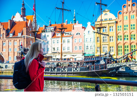 Tourist in red jacket with backpack walks along Gdansk waterfront. Behind her are historical buildings with vibrant facades and a traditional sailing ship at the harbor. 132503151