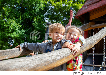 Two cheerful children enjoy sunny day at wooden playground surrounded by green trees. Laughter and bright colors create joyful atmosphere of friendship and childhood happiness Two cheerful children enjoy sunny day at wooden playground surrounded by green trees. Laughter and bright colors create joyful atmosphere of friendship and childhood happiness 132503540