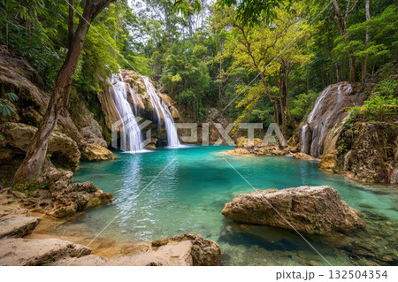 A stunning jungle waterfall cascades into a vibrant turquoise pool surrounded by lush green foliage and large brown rocks, creating a secluded natural oasis. 132504354