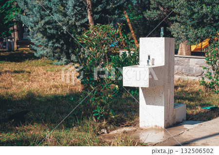 Water fountain stands in a sunny park surrounded by greenery and grass near a walking path 132506502