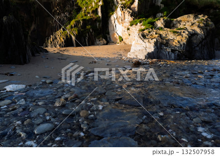 Water rushes over rocks at low tide as it flows toward a sandy cove surrounded by rocky cliffs. The scene is captured in West Cork, Ireland. 132507958