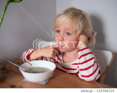 Blonde 4 year old girl touching soup with her finger, refusing to eat 132508124