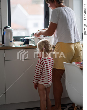 Mother trying to cook breakfast and wash the dishes while her daughter stands nearby  132508153