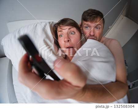 Couple lying in bed looking at a phone with shock and concern after unexpected bad news Couple lying in bed looking at a phone with shock and concern after unexpected bad news 132508304