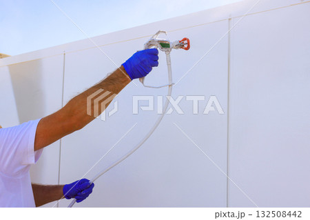 Worker with gloves applies paint to white wall using paint sprayer gun Worker with gloves applies paint to white wall using paint sprayer gun 132508442