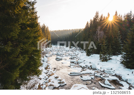 Snow-covered banks line the calm Jizera River during a winter evening. Sunlight shines through evergreen trees, creating a peaceful atmosphere at the Czechia and Poland border. Snow-covered banks line the calm Jizera River during a winter evening. Sunlight shines through evergreen trees, creating a peaceful atmosphere at the Czechia and Poland border. 132508655