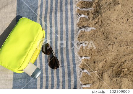 Flat lay with a makeup bag, SPF cream, and dark sunglasses on a beach towel at the beach. 132509009