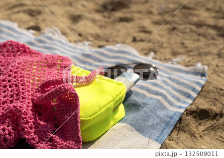 Pink mesh bag, neon makeup bag with SPF cream and sunglasses on a beach towel on the sand. 132509011