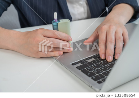 Business woman smoking an electronic cigarette while working on a laptop. Tobacco heating system. Business woman smoking an electronic cigarette while working on a laptop. Tobacco heating system. 132509654