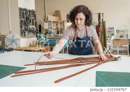 Caucasian woman makes belts from genuine leather in a workshop. Caucasian woman makes belts from genuine leather in a workshop. 132509767