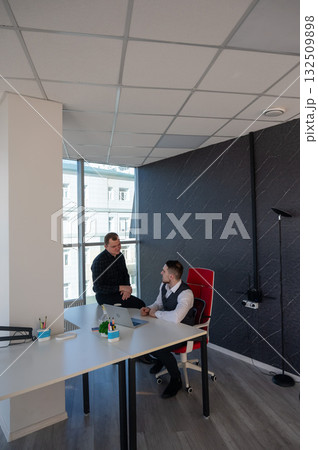 Caucasian man standing at colleague's desk discussing work.  132509898