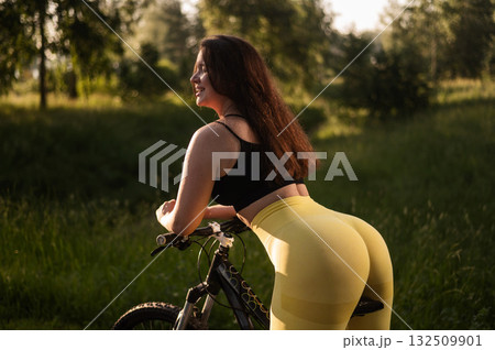 A Caucasian woman in yellow leggings and a crop top rides a bicycle in tights in the park.  132509901