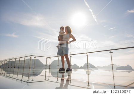 Caucasian woman holds her little son in her arms while standing on a glass bridge in the mountains. Caucasian woman holds her little son in her arms while standing on a glass bridge in the mountains. 132510077