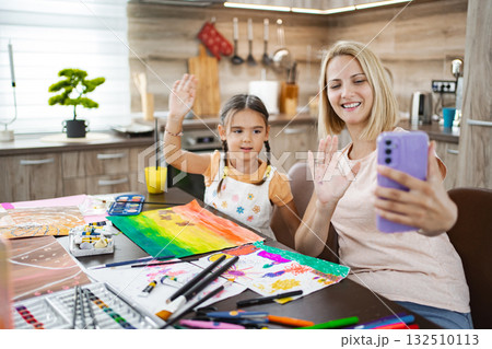 Mother and daughter video chatting while creating art in a cozy kitchen setting 132510113