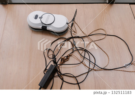 White window cleaning robot positioned on floor, resting beside charger and safety rope, preparing for efficient window cleaning tasks with advanced technology and automation at home, nobody. White window cleaning robot positioned on floor, resting beside charger and safety rope, preparing for efficient window cleaning tasks with advanced technology and automation at home, nobody. 132511183