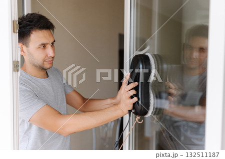 Happy young man installing robotic vacuum cleaner on window, preparing to activate device for efficient cleaning session, ensuring spotless interior environment. Cleaning house with smart devices. Happy young man installing robotic vacuum cleaner on window, preparing to activate device for efficient cleaning session, ensuring spotless interior environment. Cleaning house with smart devices. 132511187