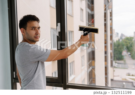 Portrait of handsome man spraying cleaning solution onto large window and wiping clean with squeegee, ensuring spotless and streak-free finish in building. Concept of cleanliness and housekeeping. Portrait of handsome man spraying cleaning solution onto large window and wiping clean with squeegee, ensuring spotless and streak-free finish in building. Concept of cleanliness and housekeeping. 132511199