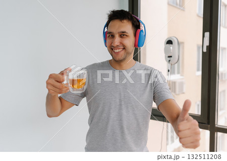 Portrait of cheerful man listening music in headphones and giving thumbs up looking at camera enjoying tea as automatic window cleaning robot works in background. Concept of household cleanliness. 132511208