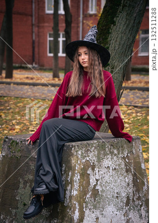 Teen girl in a witch hat and burgundy sweater sits on a stone surface surrounded by autumn leaves and trees near brick buildings 132511288